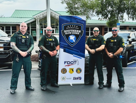 Four law enforcement officers stand in front of a banner and patrol vehicles.