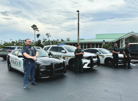 Three police officers stand alongside two marked patrol cars in a parking lot.