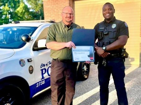 Two men, one in uniform, holding a certificate near a police vehicle.