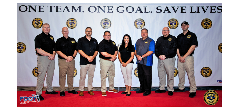 Group of seven individuals in matching shirts, standing in front of a banner that reads "ONE TEAM. ONE GOAL. SAVE LIVES."