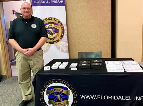 A man stands beside a table displaying informational materials and a state program banner.